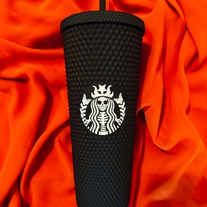 May include: Black reusable tumbler with a textured, studded exterior. The tumbler features a white logo of a skeleton with a crown on its head.