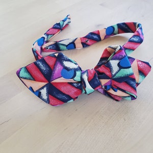 May include: A colorful bow tie with a geometric pattern in shades of pink, blue, green, and black.