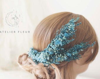 May include: A floral hair accessory featuring small, blue flowers arranged in a decorative pattern. The accessory is worn on the back of the head, with the text "L'ATELIER FLEUR" visible in the background.