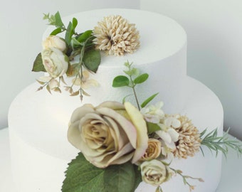 May include: Two-tiered white cake dummy decorated with a cluster of beige and cream artificial flowers, including roses and a small bouquet of greenery.