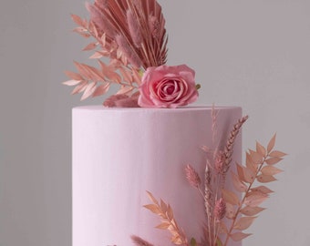 May include: A single-tier, light pink cake decorated with a pink rose and dried flowers. The cake is on a white, decorative stand. The cake has a smooth finish and is adorned with a floral arrangement. The cake is approximately 15 cm in diameter.