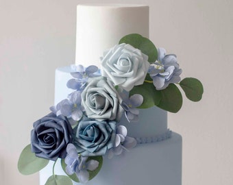 May include: A three-tiered wedding cake decorated with blue and white fondant. The cake is adorned with a cascade of blue roses and greenery.