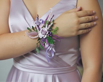 May include: A wrist corsage featuring a cluster of lavender and purple flowers, accented with green foliage and delicate ribbons. The floral arrangement includes roses and other blooms, creating a soft, romantic aesthetic. The corsage is attached to a lavender dress.
