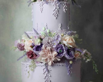 May include: A three-tiered lilac cake adorned with floral arrangements in pink, purple, and white. The cake has delicate lace-like details and sits on a decorative stand. The image includes the text "L'atelier FLEUR".