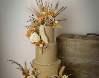 May include: A three-tiered brown wedding cake decorated with dried flowers and grasses. The cake is on a gold cake stand and is in front of a wooden suitcase.