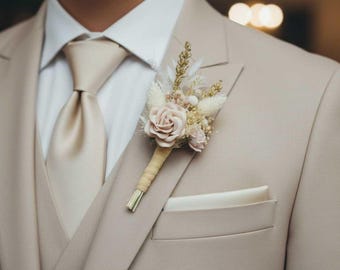 May include: A beige suit with a matching tie and a white dress shirt. A floral boutonniere with a rose and other dried flowers is pinned to the lapel. A pocket square is visible in the suit's breast pocket.