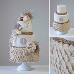 May include: A three-tier wedding cake decorated with a brown, textured frosting and white flowers. The cake is on a white cake stand. The top tier of the cake has a brown ribbon and the number "1" written in white.