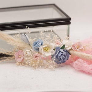 以下が含まれることがあります： ピンク、白、青の花、乾燥したパンパスグラス、白いベビーブレスを使った繊細なフラワーアレンジメント。アレンジメントはピンクのレース生地と黒いフレームのガラスボックスに飾られています。