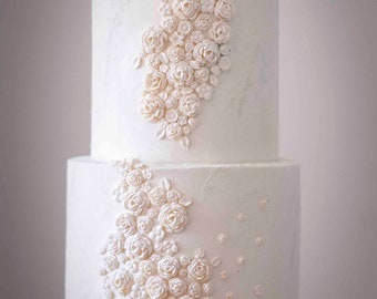 May include: A two-tiered white wedding cake decorated with delicate white sugar flowers. The cake is on a white cake stand.