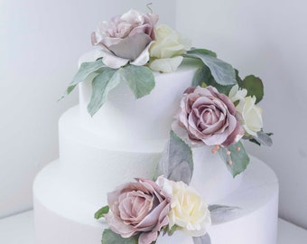 May include: A three-tiered white cake dummy decorated with artificial flowers. The flowers are a mix of dusty rose and cream roses with green leaves.