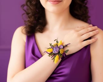 May include: A wrist corsage featuring a cluster of purple, yellow, and green flowers. The floral arrangement includes a central purple bloom, surrounded by yellow accents and green foliage. The corsage is set against a purple background.