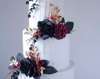 May include: A three-tiered white wedding cake decorated with black and burgundy roses and gold leaf accents.