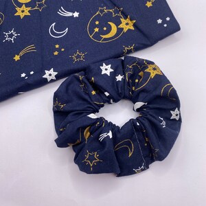 Puede incluir: Un scrunchie azul marino con un estampado celestial dorado y blanco con estrellas, lunas y estrellas fugaces.