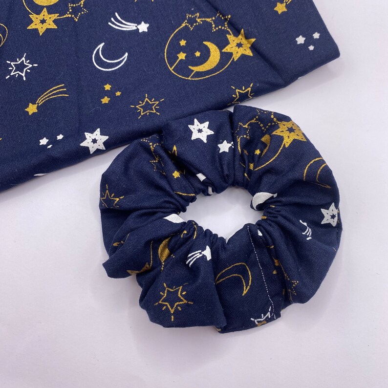 Puede incluir: Un scrunchie azul marino con un estampado celestial dorado y blanco con estrellas, lunas y estrellas fugaces.
