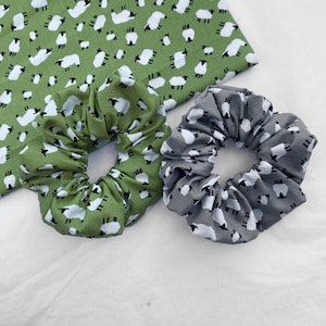 May include: Two fabric hair scrunchies with a sheep print. One scrunchie is green with white sheep, the other is gray with white sheep.