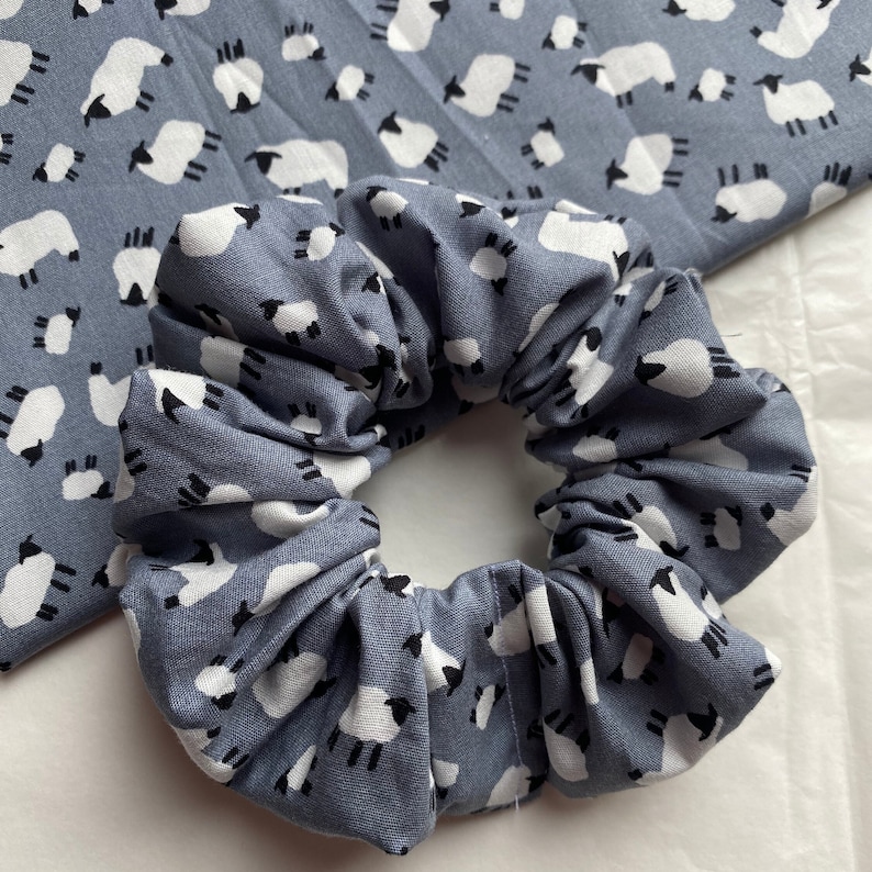 Puede incluir: Un scrunchie azul con un patr&oacute;n de ovejas blancas. El scrunchie est&aacute; hecho de tela y est&aacute; reunido en un c&iacute;rculo.