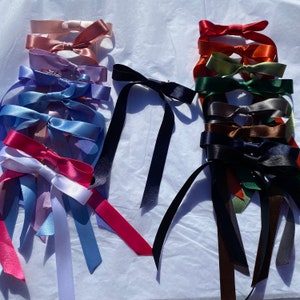 Puede incluir: Una colección de lazos de cinta de satén de colores en varios tonos de azul, rojo, verde y marrón. Los lazos están dispuestos en dos grupos, uno con lazos azules, rojos y blancos y otro con lazos multicolores.