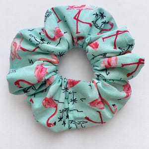 May include: A fabric hair scrunchie in a light mint green colour featuring a repeating pattern of bright pink flamingos and small black palm tree silhouettes. The accessory has a gathered, ruffled texture with a circular shape and an elastic centre.