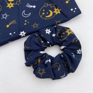 May include: A navy blue fabric scrunchie and a matching piece of fabric featuring a celestial pattern of gold and white stars, crescent moons, and shooting stars. The fabric has a smooth texture and the scrunchie is gathered in a circular shape.