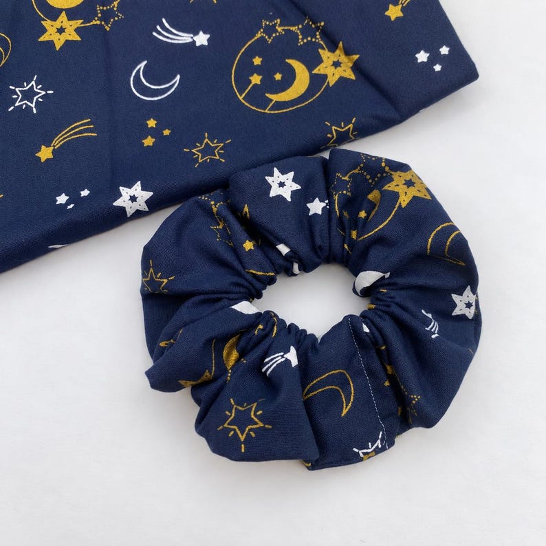 May include: A navy blue fabric scrunchie and a matching piece of fabric featuring a celestial pattern of gold and white stars, crescent moons, and shooting stars. The fabric has a smooth texture and the scrunchie is gathered in a circular shape.