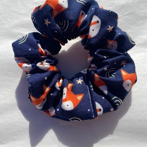 May include: A navy blue scrunchie with a repeating pattern of orange foxes and white stars on a dark blue background.
