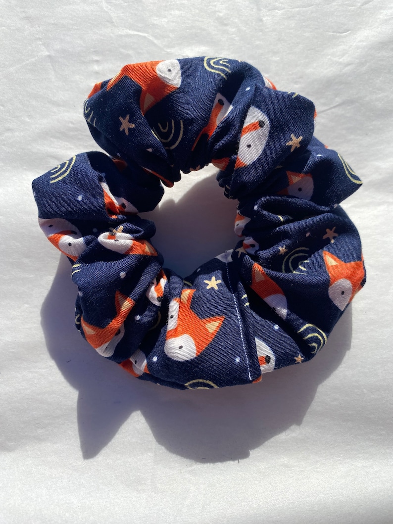 May include: A navy blue scrunchie with a repeating pattern of orange foxes and white stars on a dark blue background.
