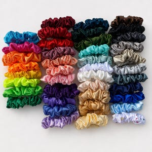 May include: A collection of numerous satin hair scrunchies arranged in columns, featuring a wide spectrum of colours including bright blue, orange, yellow, green, red, purple, pink, white, gold, silver, black, and a white scrunchie with small stars.