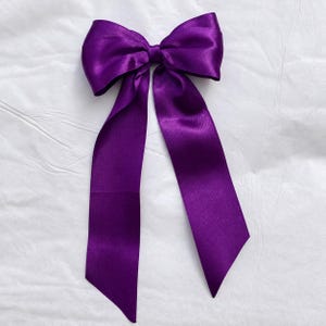 Op de afbeelding: Een dieppaarse satijnen strik met twee grote lussen en twee lange, brede linten die naar beneden hangen. De stof heeft een gladde, reflecterende afwerking met een subtiele glans, wat zorgt voor een klassieke strikvorm met strakke randen.