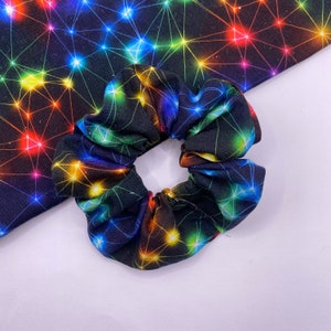 Può includere: Un elastico per capelli nero con un motivo a costellazione colorato. Il motivo presenta una rete di linee e stelle luminose e brillanti in vari colori, tra cui rosso, arancione, giallo, verde, blu e viola.