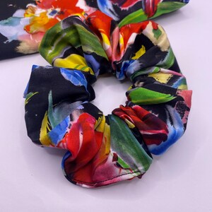 May include: A vibrant scrunchie with a floral design. The scrunchie has a black base with bright red, yellow, blue, and green accents. The fabric is gathered and elasticated, designed for tying up hair.