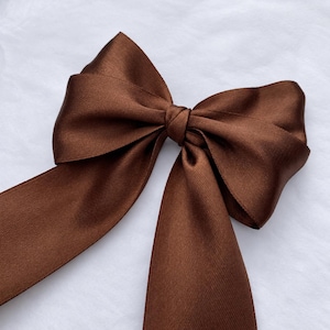 May include: A dark brown satin ribbon tied into a large, structured bow. The fabric has a smooth, reflective finish with visible horizontal weave textures. The bow features two wide, long tails extending downwards and two symmetrical loops at the top.