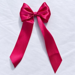 Op de afbeelding: Een fel fuchsia roze haarstrik gemaakt van glad, glanzend satijnen lint. Het accessoire heeft een centrale knoop met twee grote, gestructureerde lussen en twee lange, vloeiende linten die naar beneden vallen voor een klassiek silhouet.