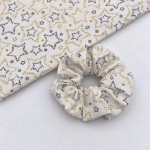 May include: A fabric scrunchie and a matching piece of fabric, both featuring an off-white background with a repeating pattern of black dotted stars and solid gold stars. The scrunchie is gathered into a circular shape with visible folds.