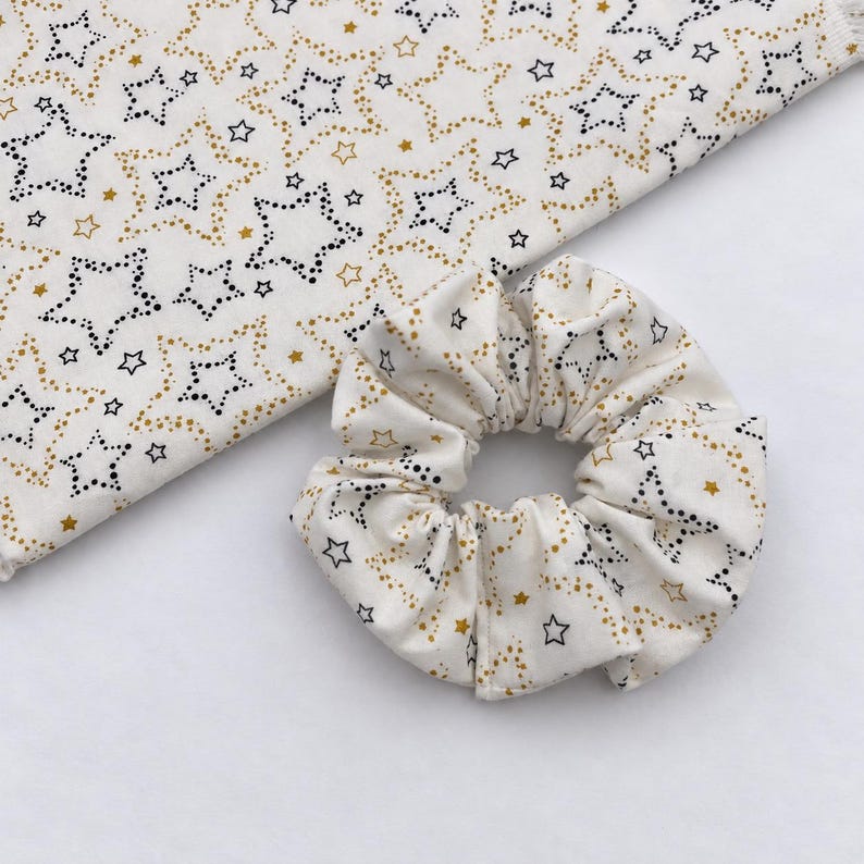 May include: A fabric scrunchie and a matching piece of fabric, both featuring an off-white background with a repeating pattern of black dotted stars and solid gold stars. The scrunchie is gathered into a circular shape with visible folds.