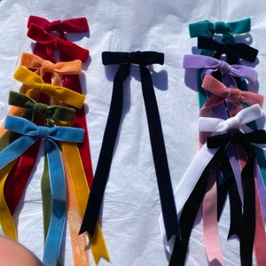 Puede incluir: Una colección de lazos de cinta de terciopelo en varios colores, incluyendo rojo, naranja, amarillo, verde, azul, marino, turquesa, morado, rosa, blanco y negro. Cada lazo tiene largas colas fluidas, perfectas para manualidades o adornos. Los lazos están dispuestos de forma atractiva.
