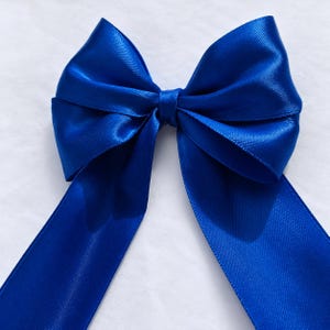 Op de afbeelding: Een levendige koningsblauwe strik gemaakt van glad, satijnachtig lint. De strik heeft twee symmetrische lussen en twee lange, vloeiende uiteinden die naar beneden hangen, met een consistente, glanzende afwerking over het gehele oppervlak.