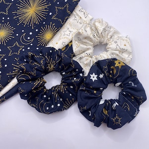 Puede incluir: Tres scrunchies para el pelo con un tema celestial. Los scrunchies est&aacute;n hechos de tela azul marino y blanca con estrellas doradas y medias lunas. Un scrunchie es blanco con estrellas doradas y medias lunas.