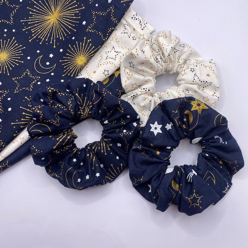 Puede incluir: Tres scrunchies para el pelo con un tema celestial. Los scrunchies est&aacute;n hechos de tela azul marino y blanca con estrellas doradas y medias lunas. Un scrunchie es blanco con estrellas doradas y medias lunas.
