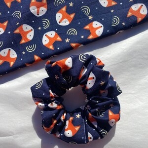 May include: A navy blue scrunchie with a repeating pattern of orange foxes and white stars. The scrunchie is made of soft, stretchy fabric.