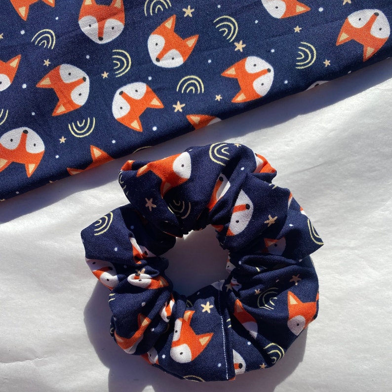 May include: A navy blue scrunchie with a repeating pattern of orange foxes and white stars. The scrunchie is made of soft, stretchy fabric.