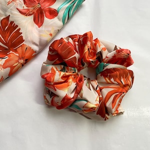 Könnte beinhalten: Ein floraler Haargummi und ein passendes Stoffstück. Der Haargummi und der Stoff haben einen cremefarbenen Hintergrund mit orangefarbenen, roten und grünen Blumenmustern. Der Haargummi ist aus einem weichen, seidigen Material gefertigt.