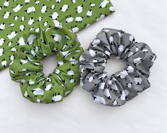 May include: Two fabric hair scrunchies featuring a repeating pattern of white sheep with black heads and legs. One scrunchie is green and the other is gray. A matching green fabric piece with the same sheep pattern is positioned behind them.