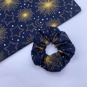 Puede incluir: Un scrunchie azul marino con un patr&oacute;n de fuegos artificiales y estrellas doradas y blancas.