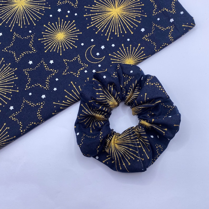 Puede incluir: Un scrunchie azul marino con un patr&oacute;n de fuegos artificiales y estrellas doradas y blancas.