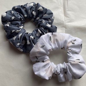 Puede incluir: Dos scrunchies de tela para el pelo. Uno es azul con un patr&oacute;n de ovejas blancas, el otro es blanco con un patr&oacute;n de ovejas grises.