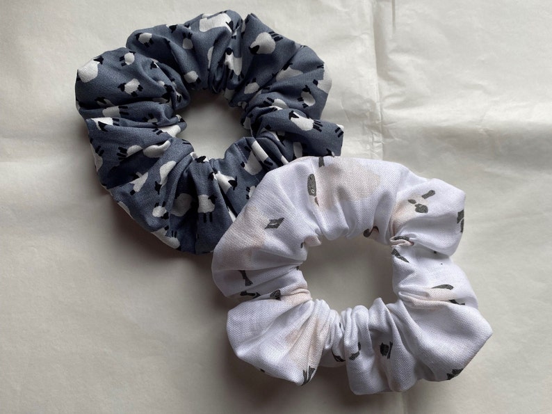 Puede incluir: Dos scrunchies de tela para el pelo. Uno es azul con un patr&oacute;n de ovejas blancas, el otro es blanco con un patr&oacute;n de ovejas grises.