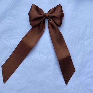 Puede incluir: Un gran lazo de satén marrón con colas largas. El lazo está atado en un estilo clásico con las colas extendiéndose hacia abajo a cada lado.