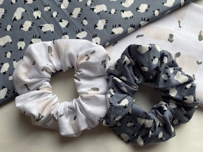 Puede incluir: Dos scrunchies de tela con estampado de ovejas. Un scrunchie es blanco con estampado de ovejas rosa claro y el otro es gris con estampado de ovejas blancas.