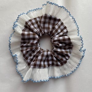 Könnte beinhalten: Ein Haargummi mit einem braun-weiß karierten Mittelteil, umgeben von weißem plissiertem Stoff mit blauem Zierstich. Dieses Haaraccessoire verleiht einen Hauch von Vintage-Charme.