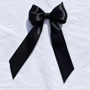 Könnte beinhalten: Eine schwarze Haarschleife aus glattem, glänzendem Satinband. Das Accessoire besteht aus einem zentralen Knoten mit zwei symmetrischen Schlaufen und zwei langen, breiten Bandenden, die nach unten hängen und eine klassische, elegante Form bilden.